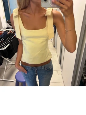 Gul zara topp - Snygg ljusgul linnetopp med breda axelband och fyrkantig ringning. Toppen har en croppad passform och är perfekt för varma dagar. Axelbanden har dekorativa rosetter som ger en söt touch. Passar till både jeans och kjol.