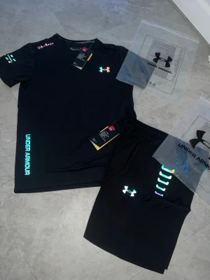 Svart träningsset från Under Armour - (Priset kan sänkas) Snyggt svart träningsset från Under Armour med t-shirt och shorts. Setet har färgglada loggor och detaljer i neonfärger samt tryckt Under Armour-text på både tröja och shorts. Materialet är lätt och andas, perfekt för träning och sport. 