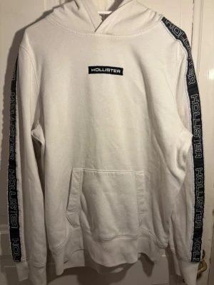 Vit hoodie från Hollister med logga - Vit hoodie från Hollister med svart broderad logga på bröstet och svarta band med Hollister-tryck längs ärmarna. Klassisk huva och stor magficka framtill. Perfekt för dig som gillar en clean och sportig look.