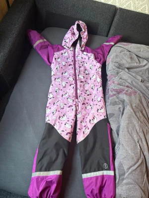 Rosa overall med enhörningsmönster - Barn overall från Ski Industries i lila och svart med gulligt enhörningsmönster och regnbågar. Byxorna har huva, dragkedja framtill och reflexdetaljer vid benen. Materialet är slitstarkt och vattenavvisande, perfekt för kalla dagar i snön.
