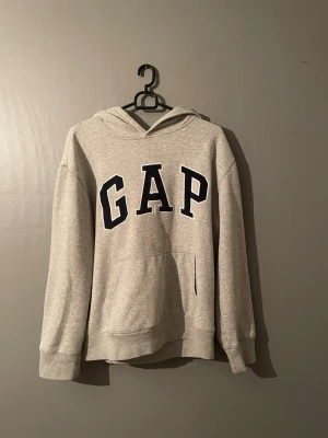 Grå hoodie från GAP med huva - Snygg grå hoodie från GAP med stor svart logotyp på bröstet. Hoodien har en klassisk huva och en stor magficka framtill. Materialet är mjukt och bekvämt, perfekt för chill dagar. Långärmad och avslappnad passform.
