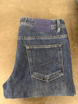 Blå raka jeans från Jacob Cohen W34 - Säljer ett par klassiska blå jeans från Jacob Cohen med raka ben och femficksdesign. Jeansen har en mörkblå patch bak och kontrastsömmar. Materialet är slitstark denim som ger en tidlös look. Perfekt för dig som gillar stilrena och bekväma jeans.