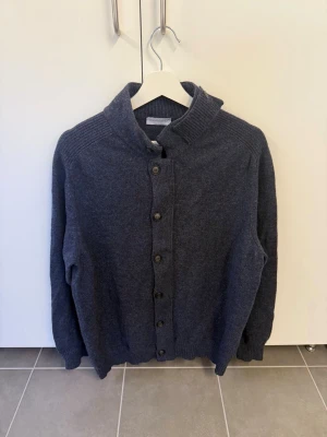 Mörkgrå kofta från Gran Sasso stl L - Snygg mörkgrå kofta från Gran Sasso, tillverkad i Italien. Koftan har en hög krage, knappar hela vägen fram och ribbade detaljer vid kragen och nederkanten. Perfekt för lager-på-lager och en stilren look.