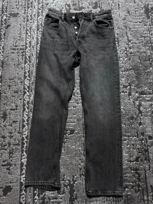 Svarta raka jeans med knappar - Säljer ett par svarta jeans med rak passform och knappgylf. Jeansen har klassiska fem fickor och en diskret läderpatch bak i midjan. Materialet är denim och färgen är tvättad svart, vilket ger en cool vintagekänsla.