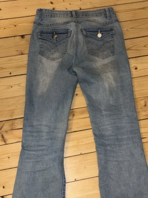 Ljusblå bootcut jeans med detaljer - Säljer ett par ljusblå bootcut jeans med snygga broderade bakfickor och silverknappar. Jeansen har en klassisk femficksdesign och är tillverkade i mjukt denim-material. Perfekt för dig som gillar en avslappnad och trendig look.