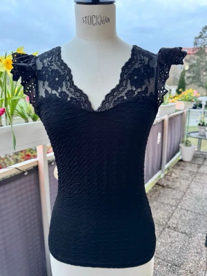 Svart topp med volangärmar - Ny Supersnygg svart topp med v-ringad spetsdetalj och blommönster upptill. Volangärmar. Figurnära passform och lätt strukturerat material 🤍