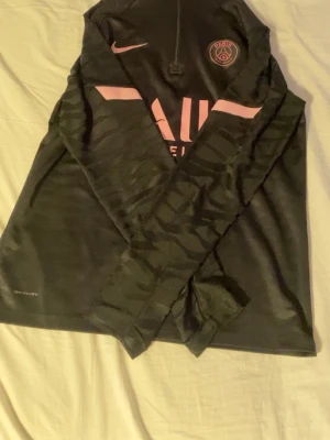 Psg tröja - Svart långärmad träningströja från Nike med PSG-logga på bröstet och rosa detaljer. Tröjan har halv dragkedja, Dri-FIT material och diskreta mönster på ärmarna. Perfekt för fotbollsträning eller gymmet.
