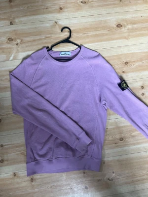 Ljuslila sweatshirt från Stone Island - Ljuslila sweatshirt från Stone Island med klassisk rund hals och ribbade muddar. Tröjan har den ikoniska Stone Island patchen på vänster ärm och är tillverkad i mjuk bomull. Perfekt för dig som vill ha en snygg och bekväm tröja med streetwear-vibe.