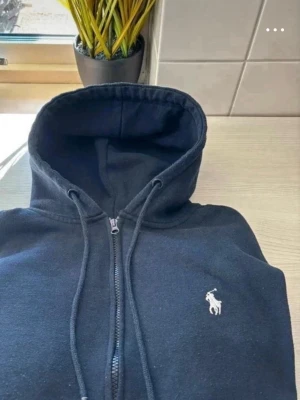 Svart hoodie med dragkedja från Polo Ralph Lauren - Säljer en svart hoodie från Polo Ralph Lauren med dragkedja och huva. Klassisk design med logotyp broderad på bröstet. Mjuk bomullskvalitet och snörning i huvan. Perfekt för en avslappnad och stilren look. 
