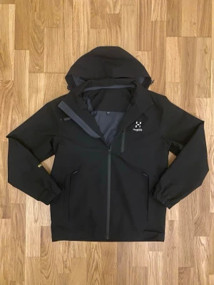 Svart regnjacka från Haglöfs - Säljer en svart regnjacka från Haglöfs med huva och hel dragkedja framtill. Jackan har två sidofickor med dragkedja och elastiska ärmslut. Materialet är vattenavvisande och perfekt för regniga dagar. Diskret Haglöfs-logga på bröstet. Står storlek L men passar S och runt 170cm