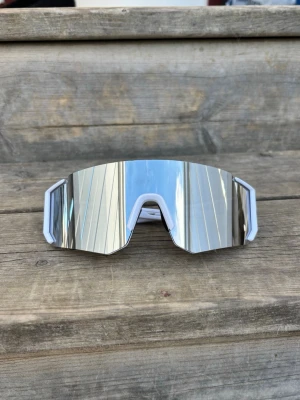 Silvriga sportiga solglasögon - Säljer ett par futuristiska solglasögon med spegelblanka silvriga linser och vit ram. De har en bred, kantig form som ger en cool och modern look. Perfekta för dig som vill sticka ut och gillar sportiga accessoarer.