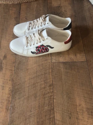 Gucci ace skor  - Säljer ett par vita sneakers från Gucci med coolt broderat orm-motiv på sidan. Skorna har snörning och detaljer i rött och grönt på hälen, Perfekt för dig som vill sticka ut med exklusiva skor. Storlek 44 BVSA