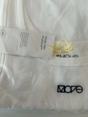 Vit ribbad linnetopp från H&M Move - Säljer en vit ribbad linnetopp från H&M Move med broderad logga framtill. Toppen är ärmlös och har en sportig design som passar perfekt till träning. Materialet är mjukt och följsamt, och modellen har rund halsringning.