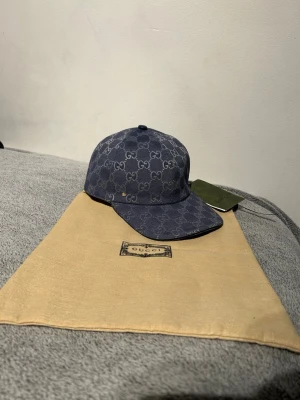 Blå Gucci keps med GG-mönster - Snygg blå keps från Gucci med det klassiska GG-mönstret över hela kepsen. Justerbar rem baktill för perfekt passform. Kepsen är tillverkad i bomull och har en stilren, modern look som passar till alla outfits.