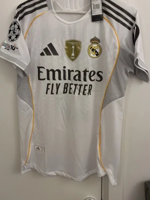 Real Madrid matchtröja Bellingham 5 - Säljer en vit Real Madrid matchtröja från Adidas med Bellingham och nummer 5 på ryggen. Tröjan har svarta och guldiga detaljer, klubbmärke, FIFA-märke och Champions League-märke på ärmen. Tillverkad i lätt och ventilerande material, perfekt för fotboll. Helt ny.