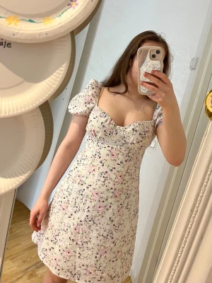 Blommig vit kortklänning med puffärm - Superfin vit kortklänning med blommigt mönster och puffärmar. Klänningen har en söt sweetheart-ringning och smockad rygg som ger en bekväm passform. Perfekt för sommarens alla tillfällen, och den är gjord i ett lätt bomullstyg.