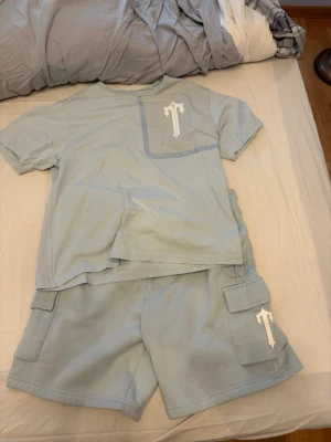 Trapstar Ljusblått set  - Säljer ett ljusblått set från Trapstar med t-shirt och shorts. Båda delarna har ett vitt tryck med ett stiliserat T på bröstfickan och shortsfickan. Setet har en avslappnad passform och är gjort i mjukt bomullsliknande material. Perfekt för en chill och bekväm stil.
