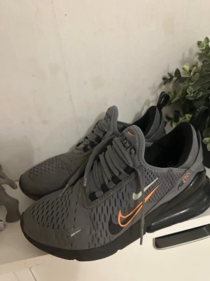 Nike Air Max 270 grå sneakers - Nike Air Max 270 sneakers i grå mesh med svarta och orange detaljer. Skorna har en stor synlig Air Max-dämpning i hälen och en låg profil. Snygg design med ventilerande material och klassisk Nike-logga på sidan.