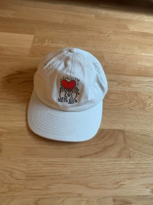 H&M Keith Haring Keps Vit Dad Cap - Snygg vit dad cap från H&M med ikoniskt Keith Haring-motiv – två figurer som håller upp ett rött hjärta. Ett coolt konst-collab som sticker ut. Justerbar i nacken, passar de flesta.