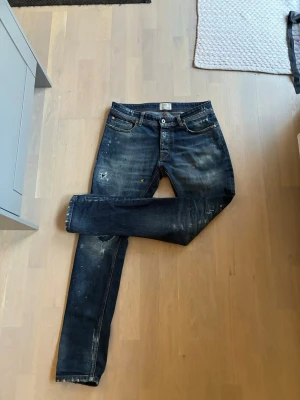 Mörkblå slitna jeans från Dsquared2 - Säljer ett par mörkblå jeans från Dsquared2 med slitningar, färgstänk och distressed detaljer. Klassisk femficksmodell med knappgylf och smal passform. Perfekta för dig som gillar en edgy och trendig stil.