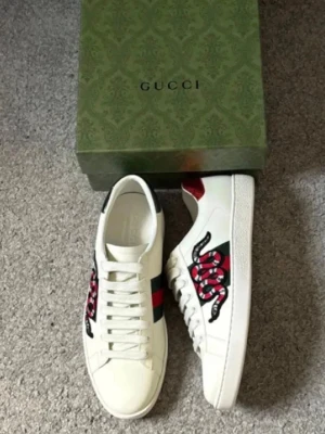 Gucci Ace sneakers  - Säljer ett par vita Gucci Ace sneakers i läder med klassiska gröna och röda sidoremsor och en broderad orm på sidan. Baksidan har metallicdetaljer i rött och grönt med Gucci-logga. Snygg och ikonisk modell som sticker ut. Obs skolåda medföljer ej