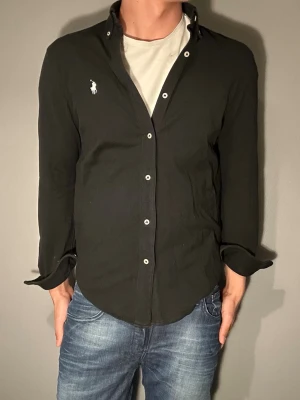 Svart skjorta från Polo Ralph Lauren - Svart skjorta från Polo Ralph Lauren med klassisk vit logobrodyr på bröstet. Skjortan har långärmad design, vita knappar och en normal passform. Materialet är bomull och skjortan har en tidlös look som funkar till många stilar.