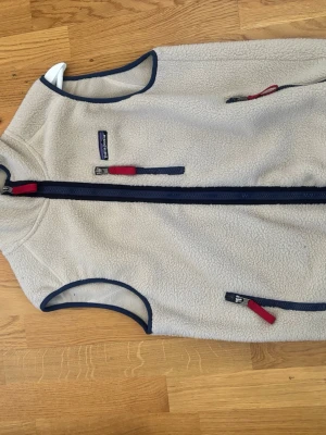 Beige fleeceväst från Patagonia - Säljer en beige fleeceväst från Patagonia med mörkblå detaljer och röda dragkedjor. Västen har två sidofickor och en bröstficka med dragkedja, samt mörkblå kantband runt ärmhål och krage. Perfekt för lager-på-lager och outdoor.