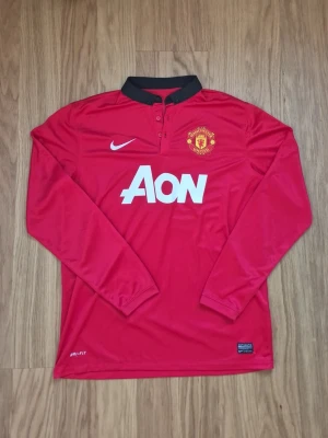  Authentic Retro Manchester United långärmad matchtröja - Säljer en röd retro långärmad Manchester United matchtröja från Nike med svart krage och knappar. Tröjan har klubbens emblem på bröstet, vit Nike-logga och Aon som sponsor. Materialet är Dri-FIT för optimal komfort vid träning och match.
