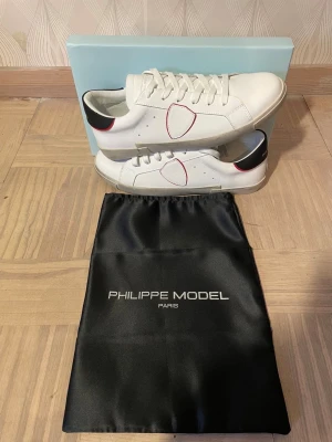 Vita Philippe Model sneakers med svart detalj - Snygga vita sneakers från Philippe Model Paris med svart hälparti och röda detaljer. Skorna har klassisk låg profil, vita snören och är tillverkade i läder. Medföljer originalpåse och box. Perfekt för dig som gillar stilrena och trendiga sneakers.
