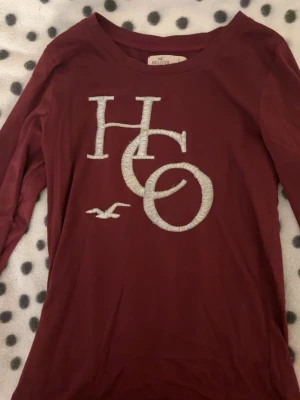 Vinröd långärmad t-shirt från Hollister - Säljer en vinröd långärmad t-shirt från Hollister med stort broderat HO-logo i vitt på bröstet. T-shirten har rund halsringning och är gjord i mjuk bomull, perfekt för en avslappnad och cool look.