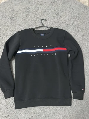 Svart sweatshirt Tommy Hilfiger - Svart sweatshirt från Tommy Hilfiger med rund halsringning och tryckt logga samt röd, vit och blå rand över bröstet. Tröjan har ribbade muddar vid ärmslut och nederkant. Perfekt för en stilren och avslappnad look. Ingen prutning! 