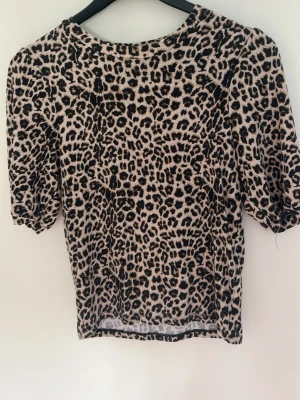 Leopardmönstrad topp med puffärm - Snygg topp med leopardmönster i beige och svart. Modellen har puffiga korta ärmar och rund halsringning. Perfekt för dig som vill sticka ut lite extra. Materialet känns mjukt och stretchigt, troligen polyester eller bomullsmix.