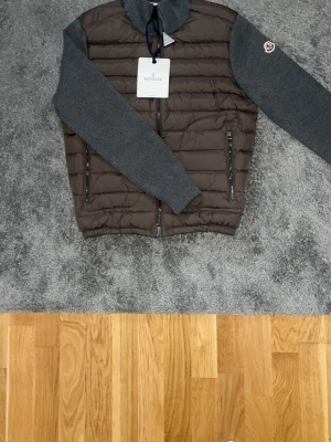 Moncler grå/brun cardiganjacka - Snygg cardiganjacka från Moncler med quiltad brun front och grå stickade ärmar och rygg. Jackan har två dragkedjeförsedda fickor fram, hög krage och Moncler-logga på vänster ärm. Perfekt för lager-på-lager och streetstyle.