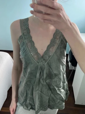 Spetstopp - Säljer en grön satintopp med djup v-ringning och breda spetskanter längs urringningen. Toppen är ärmlös och har en lös passform med lätt skrynklig look. Perfekt för en avslappnad och trendig stil.