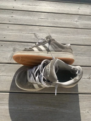 Grå Adidas Campus sneakers - Säljer ett par grå Adidas Campus sneakers med klassiska vita ränder på sidan och gummisula i ljusbrunt. Skorna har en låg profil och är tillverkade i mocka med vita skosnören och Adidas-logga på plösen. Perfekta för en avslappnad streetwear-look.