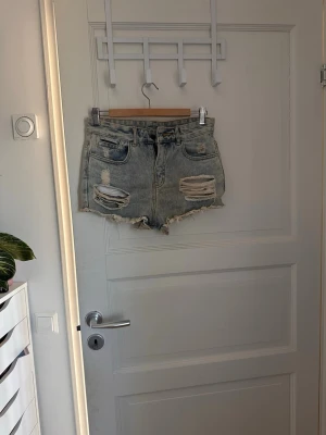 Ljusblå slitna jeansshorts - Säljer ett par ljusblå jeansshorts med råa kanter och stora slitningar framtill. Shortsen har fem fickor och klassisk knappgylf. Perfekta för en avslappnad och trendig sommarlook.