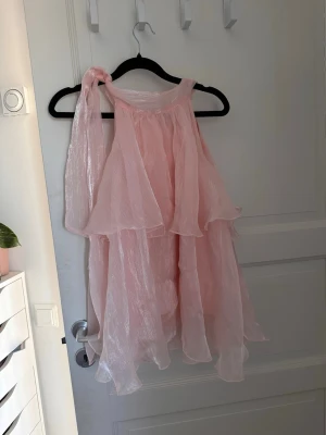 Ljusrosa volangklänning axelbandslös - Säljer en drömmig ljusrosa klänning med flera lager volanger och axelbandslös design. Klänningen har en dekorativ knytning på ena axeln och är gjord i ett skirt, glansigt material som ger ett romantiskt intryck. Perfekt för sommarens alla tillfällen.