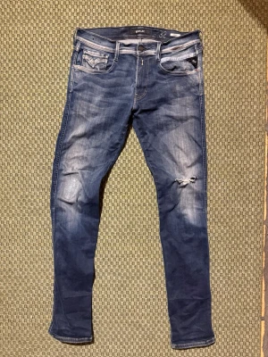 Replay - Säljer ett par blå jeans från Replay med slim fit och slitningar på ena benet. Jeansen har klassiska fem fickor, dragkedjegylf och snygga detaljer vid fickorna. Materialet är denim med en tvättad look och stretch för skön passform.