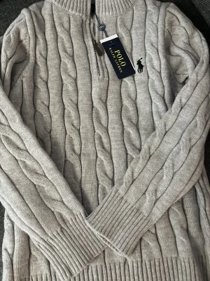 Polo Ralph Lauren zip up - Kabelstickad tröja i ljusgrått från Polo Ralph Lauren med halv dragkedja och ribbade muddar. Klassisk broderad logga på bröstet och hög krage. Perfekt för dig som gillar stilrena och tidlösa plagg.