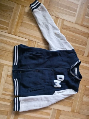 Mörkblå bomberjacka med vita ärmar - Snygg mörkblå bomberjacka med vita ärmar och broderade bokstäver 'DM' på bröstet. Jackan har randiga muddar i blått och vitt vid ärmslut, krage och nederkant samt två fickor framtill. Stängs med svarta tryckknappar. Perfekt för en sportig och avslappnad stil. Storlek 110/116.