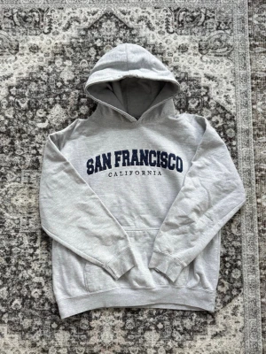 Grå vintage hoodie med San Francisco-tryck - Grå vintage hoodie med stor huva och känguruficka framtill. På bröstet står det 'San Francisco California' i mörkblå text. Hoodien har ribbade muddar vid ärmslut och nederkant. För mer info är de bara att skriva!