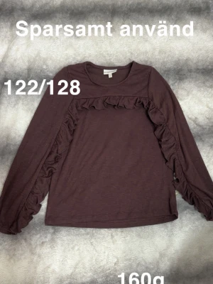  brun långärmad topp med volang - Långärmad topp från pompdelux i brun färg med dekorativa volanger längs framsidan och ärmarna. Perfekt för dig som gillar en söt och trendig look.🌸 Sparsamt använd. Storlek 122/128