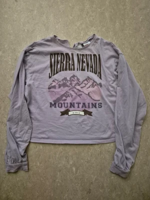 Tröja - Lila långärmad tröja med tryck av berg och texten 'Sierra Nevada Mountains' framtill. Tröjan har en avslappnad passform, rund halsringning med liten v-formad detalj och mjukt bomullsliknande material. Perfekt för en chill och trendig look.
