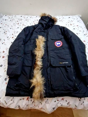 Svart parkas från Canada Goose med extra ny päls Storlek M passar L - Säljer en svart parkas från Canada Goose med klassisk röd logotyp på bröstet och stor huva med avtagbar pälskant. Jackan har flera praktiska fickor och dragkedja framtill. Perfekt för kalla vinterdagar och riktigt snygg streetstil.