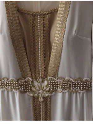 Vit och guld långklänning med pärlor - Elegant långklänning i vitt med detaljer i guld och pärlor. Klänningen har djup v-ringning, bred midjedekor med pärlor och broderade kanter. Materialet ser ut att vara lätt och flytande, perfekt för speciella tillfällen.