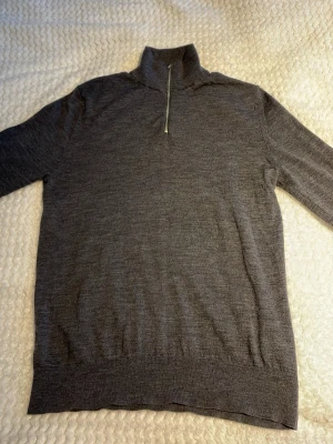 Grå stickad tröja med halvzip Riley - En stilren grå stickad tröja med halvzip och hög krage. Tröjan har långa ärmar och ribbstickade muddar vid ärmslut och nederkant. Materialet ser ut att vara mjukt och tunt, perfekt för lager-på-lager. Passar dig som gillar enkel och klassisk design.