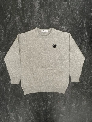 Grå stickad tröja Comme des Garçons - (Kommer även med originala påsen och tags 🏷️) Grå stickad tröja från Comme des Garçons PLAY med svart hjärta och ögon broderat på bröstet. Tröjan har rund halsringning, ribbade muddar och är i mjukt material. Perfekt för en clean och stilren look.