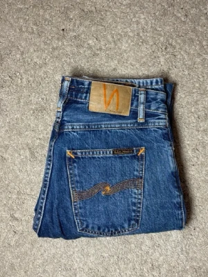 Blå jeans från Nudie Jeans - Säljer ett par klassiska blå jeans från Nudie Jeans med snygga kontrastsömmar och ikonisk orange logga på bakfickan. Jeansen har rak passform och är tillverkade i slitstark denim. Perfekta för dig som gillar tidlös stil och kvalitetsjeans.