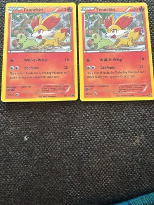 Två Fennekin Pokémonkort - Säljer två identiska Fennekin Pokémonkort med gul ram och röd-orange botten. Motivet visar Fennekin tillsammans med ett grönt Pokémon i bakgrunden. Kortet har attackerna Will-O-Wisp och Confront samt detaljerad text på engelska.
