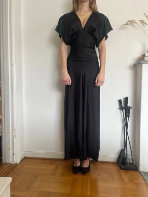 Svart långklänning med snörning - Aldrig använd med lappar kvar, helt slutsåld på hemsidan! Elegant svart långklänning med v-ringad front och korta, vida ärmar. Klänningen har snörning i ryggen som ger en snygg passform och är tillverkad i ett glansigt material som faller fint. Perfekt som balklänning eller andra finare tillställningar! 